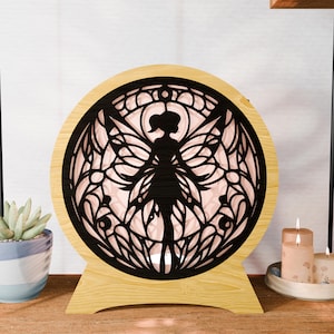 May include: A round wooden lamp with a black fairy silhouette design. The fairy has detailed wings and is surrounded by an intricate floral pattern. The lamp has a light pink glow and sits on a wooden base.