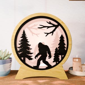 May include: A wooden Bigfoot-themed night light. The round light features a black silhouette of Bigfoot walking through a forest scene with trees and branches. The light has a wooden base and frame, with a soft pink glow.