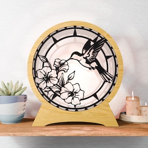 Op de afbeelding: Ronde decoratieve houten lamp met een zwart kolibrie- en bloemmotief. De lamp heeft een lichtgekleurde houten basis en frame, met een witte achtergrond die het uitgesneden ontwerp verlicht. De lamp staat op een houten plank met een kleine vetplant en kaarsen.