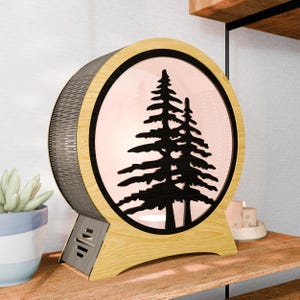 May include: A round, wooden lamp with a black silhouette of two pine trees. The lamp has a light wood frame and base, with a dark, textured back. The center glows with a soft, pinkish light. The lamp is on a wooden surface.