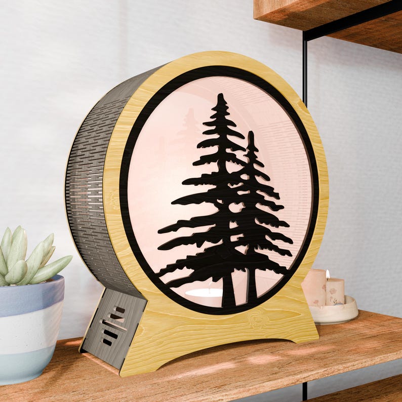 May include: A round, wooden lamp with a black silhouette of two pine trees. The lamp has a light wood frame and base, with a dark, textured back. The center glows with a soft, pinkish light. The lamp is on a wooden surface.