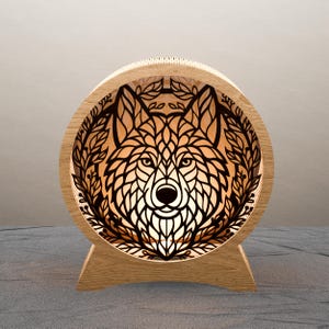 May include: A wooden, circular lamp featuring a detailed, laser-cut wolf design. The wolf is surrounded by a leafy border. The lamp has a warm, inviting glow, and is set on a wooden base.