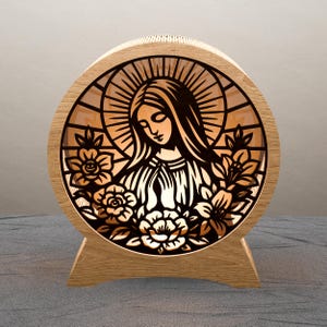 May include: A circular wooden religious artwork with a laser-cut design of a woman with a halo and floral elements. The interior is illuminated, producing a warm light. The piece is supported by a wooden stand. The diameter is approximately 20 cm.