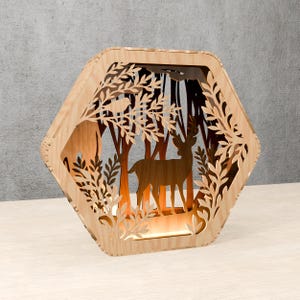 May include: A wooden decorative lamp with a forest scene. The lamp features a deer, trees, and foliage cut into the wood. The lamp has a warm light inside, creating a cozy ambiance. The lamp is in a hexagonal shape.