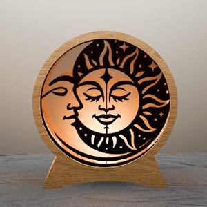 May include: A wooden celestial-themed lamp featuring a sun and moon design. The sun has a smiling face with closed eyes, and the moon is in profile. The lamp has a round shape with a wooden base and frame, and the light shines through the cut-out design.