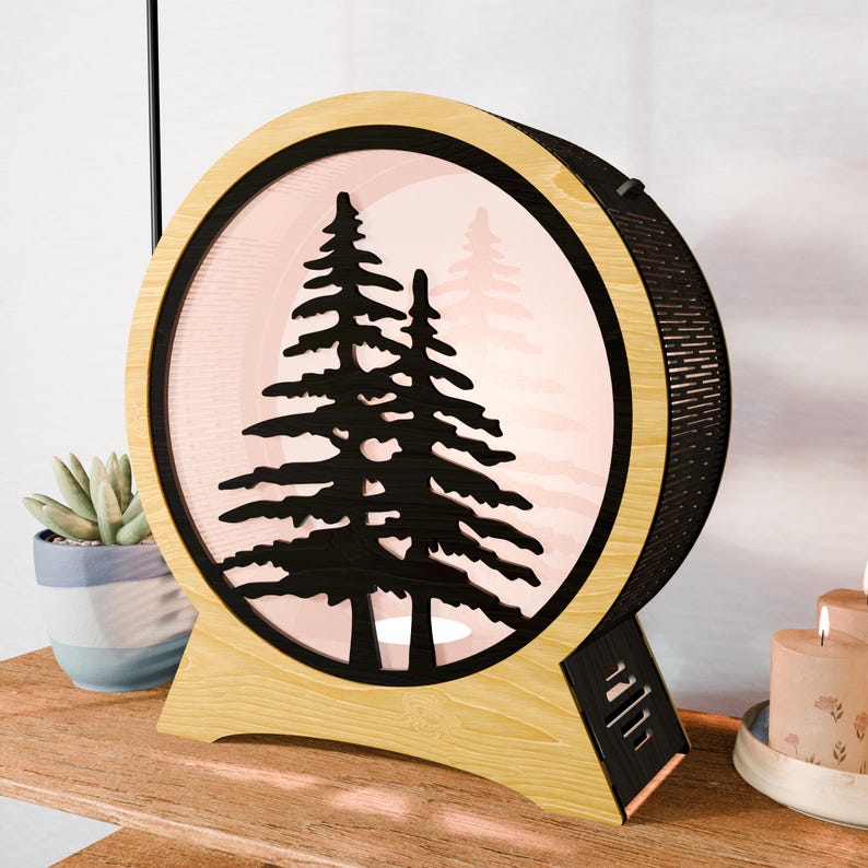 May include: A round, wooden lamp with a black silhouette of pine trees inside. The lamp has a light pink background and a black mesh back. The base is light wood, and the lamp is sitting on a wooden surface.