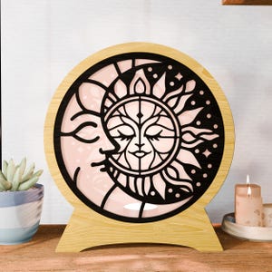 May include: A wooden, round decorative light featuring a sun and moon design. The sun and moon are black, with a pink background. The light has a wooden base and frame, and is suitable for home decor.