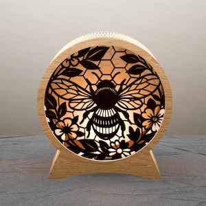 Può includere: Una lampada rotonda in legno con un design a forma di ape tagliato al laser. La lampada presenta una silhouette dettagliata di un'ape circondata da motivi floreali e a nido d'ape. La luce calda brilla attraverso i ritagli, creando un pezzo decorativo e artistico.