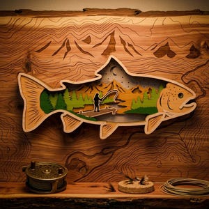May include: A wooden layered art piece in the shape of a fish, featuring a fishing scene inside. The fish is light brown with a detailed design. The interior shows a person fishing on a dock, with mountains and trees in the background.