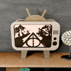 May include: A wooden decorative television with a light-up screen featuring a silhouette scene. The TV has a light wood frame, dark wood screen details, and a pair of antennas. The screen displays a scene with figures and a triangle symbol.