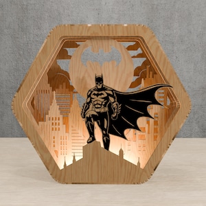May include: A wooden, hexagonal Batman-themed night light. The design features a silhouette of Batman standing on a rooftop, with the city skyline and the bat symbol in the background. The light is made of layered wood.
