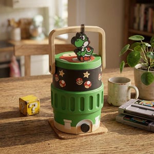 May include: A cylindrical green and black cooler with a light wood handle and a Yoshi figure on top. The black section features Mario, mushroom, and star icons. The green base has a white rectangular detail and a black circular spout at the bottom.