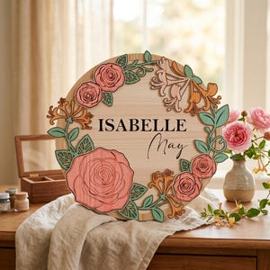 May include: A circular wooden sign featuring the name Isabelle May in black lettering. The sign is decorated with layered, cut-out floral elements in shades of pink, orange, and green, arranged in a wreath pattern around the central text.