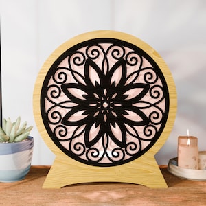 Pode incluir: Lâmpada redonda de madeira com um design floral preto recortado. A lâmpada tem um fundo rosa claro e uma base de madeira. Uma planta suculenta num vaso azul e uma vela acesa estão no fundo.