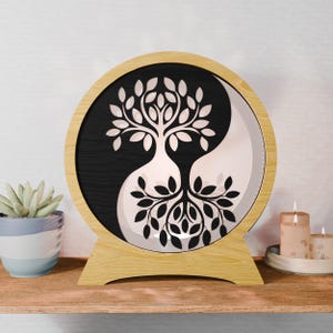 May include: A round wooden Yin Yang wall art piece with a tree design. The artwork features a black and white Yin Yang symbol with a tree silhouette in the center. The piece is displayed on a wooden shelf with a succulent and candles.