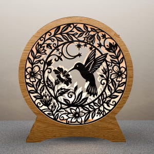 May include: A wooden, round, illuminated hummingbird lamp. The lamp features a black hummingbird silhouette, flowers, leaves, a crescent moon, and stars. The lamp is made of wood and has a light-colored interior. The lamp is on a wooden base.