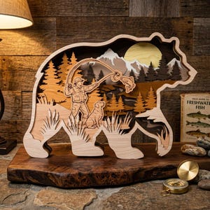 Könnte beinhalten: Ein hölzernes, bärenförmiges Kunstwerk, das einen Fischer beim Angeln eines Fisches zeigt, mit einem Hund, Bäumen, Bergen und einer Sonne. Das mehrschichtige Design verwendet helle und dunkle Holztöne für einen 3D-Effekt. Ein Buch mit dem Titel "Field Guide to Freshwater Fish" ist sichtbar.
