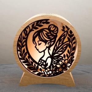 Peut inclure: Lampe en bois en forme de fée avec un design circulaire. La lampe présente une silhouette de fée avec des ailes détaillées et des motifs floraux. La fée est entourée d'un motif découpé dans le cadre en bois, éclairé par une lumière chaude.