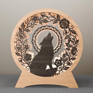 May include: A wooden, circular decorative light featuring a howling wolf silhouette. The wolf is centered within a floral and leaf design, with a light gray background. The light has a wooden base and frame.