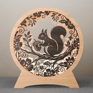 May include: A circular wooden light fixture with a laser-cut design of a squirrel holding an acorn on a branch. The design is set against a grey background, framed by floral and leaf cutouts. The fixture has a natural wood finish.
