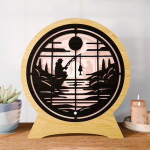 May include: A wooden, round lamp with a black silhouette of a fisherman on a lake. The lamp has a light-colored wooden frame and a black, laser-cut design. The silhouette shows a fisherman fishing, with a fish on the line. The lamp is a decorative item.