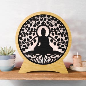 Pode incluir: Lâmpada de silhueta de Buda em madeira com um design recortado em preto de uma árvore e raízes. A lâmpada tem formato redondo com moldura e base marrom claro. O Buda está em posição de meditação sentado.