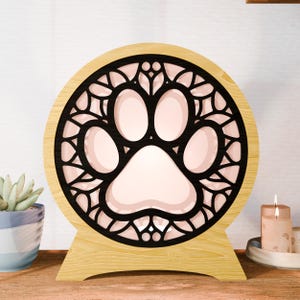 Puede incluir: Una luz de noche de huella de pata de madera. La luz tiene un diseño circular con una huella de pata negra en el centro, rodeada por un borde negro decorativo. La base y el marco son de madera clara.