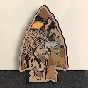 May include: A wooden cutout of a Native American woman wearing a headdress and holding a spear. The background features a sunset, a teepee, and a totem pole.
