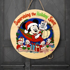 May include: Round wooden sign featuring Mickey Mouse in a Santa hat, holding a clipboard that says "Nice List List." The sign reads "Supervising the Holiday Spirit." Other cartoon characters are also present, surrounded by wrapped gifts.