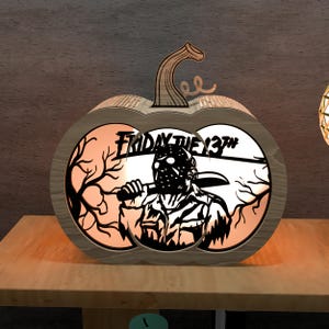 Op de afbeelding: Houten pompoenvormige Halloween-decoratie met de tekst "Friday the 13th" en een silhouet van Jason Voorhees. Het ontwerp wordt van binnenuit verlicht, wat een spookachtig effect creëert. De pompoen heeft een houten steel.