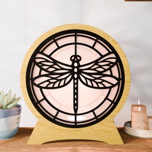 May include: A decorative wooden lamp with a black dragonfly design. The lamp has a circular frame with a light pink center and a wooden base. The dragonfly is cut out of the black frame. A small succulent plant and candles are in the background.