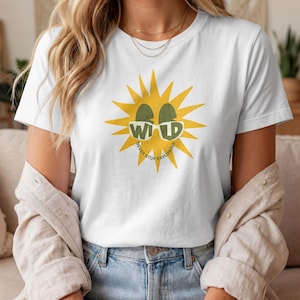 Puede incluir: Una camiseta blanca de manga corta con un gráfico de sol amarillo y gafas de sol verdes. Las gafas contienen el texto WILD, y el texto NEVER STOP EXPLORING está impreso en un arco circular debajo del gráfico del sol en la parte delantera.