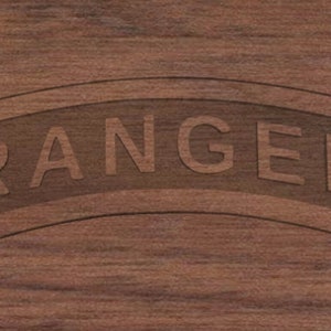 May include: A close-up of a wooden surface with the word "RANGER" engraved in a curved banner. The text and banner are a darker shade than the surrounding wood, creating a subtle contrast. The wood grain is visible.