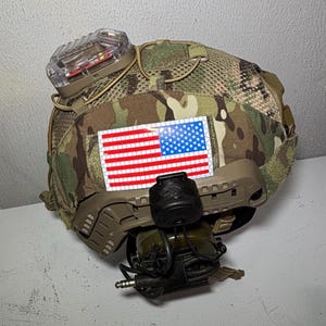 Puede incluir: Un casco táctico de camuflaje con un parche de la bandera estadounidense, una luz clara y un auricular de comunicación. El casco tiene una parte superior de malla y varios accesorios. Los colores incluyen verde, marrón, beige, rojo, blanco y azul.