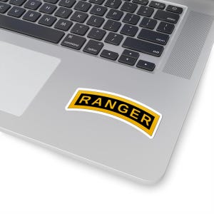 May include: A white sticker with the word "RANGER" in bold, black letters. The text is set against a yellow background with a black border. The sticker is slightly curved and placed on a silver laptop.