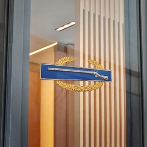 May include: A blue rectangular sign with a silver rifle emblem, framed by a gold laurel wreath. The sign is mounted on a glass door, with a wooden slatted wall in the background. The overall design is classic and elegant.