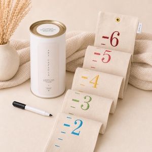 May include: A white cylindrical storage container labelled Art Capsula with a growth chart made of off-white fabric featuring embroidered numbers two through six in blue, green, yellow, and red, accompanied by a black marker for recording heights.
