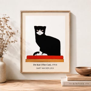 May include: A framed art print featuring a black cat with white paws and a white face sitting on a red rectangular cushion. The text below the illustration reads: De Kat (The Cat), 1914, BART VAN DER LECK. The print is set in a light wood frame.