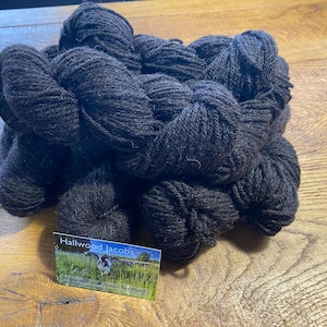 Peut inclure: Plusieurs écheveaux de fil brun foncé, étroitement enroulés et empilés, reposent sur une surface en bois. Une carte de visite avec le texte "Hallwood Jacobs" et une scène pastorale est visible.