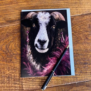 May include: Greeting card featuring a black sheep with white face and pink feather accents. The sheep wears pink jeweled earrings. A black pen with gold accents rests on the card, which is on a wooden surface.