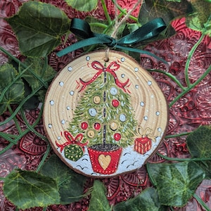 May include: A round wooden slice ornament featuring a hand-painted green Christmas tree in a red pot with a gold heart. The tree is decorated with red and silver ornaments, topped with a red bow, and surrounded by two gift boxes and white snow.