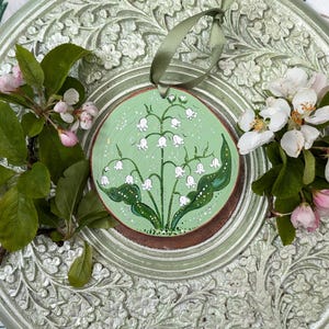 May include: A round, hand-painted wooden ornament featuring a lily of the valley design on a light green background. The ornament is accented with a green ribbon for hanging. The ornament is placed on a decorative, light green, floral-patterned plate, with sprigs of flowers and leaves.