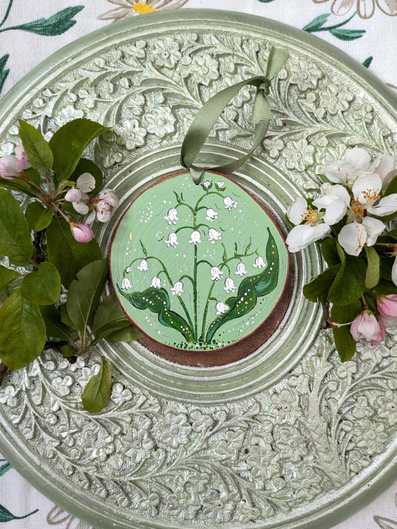May include: A round, hand-painted wooden ornament featuring a lily of the valley design on a light green background. The ornament is accented with a green ribbon for hanging. The ornament is placed on a decorative, light green, floral-patterned plate, with sprigs of flowers and leaves.