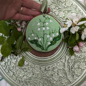 May include: A round, wooden ornament with a hand-painted lily of the valley design. The ornament features a light green background with white flowers and green leaves. A green ribbon is attached for hanging. The ornament is approximately 4 inches in diameter.