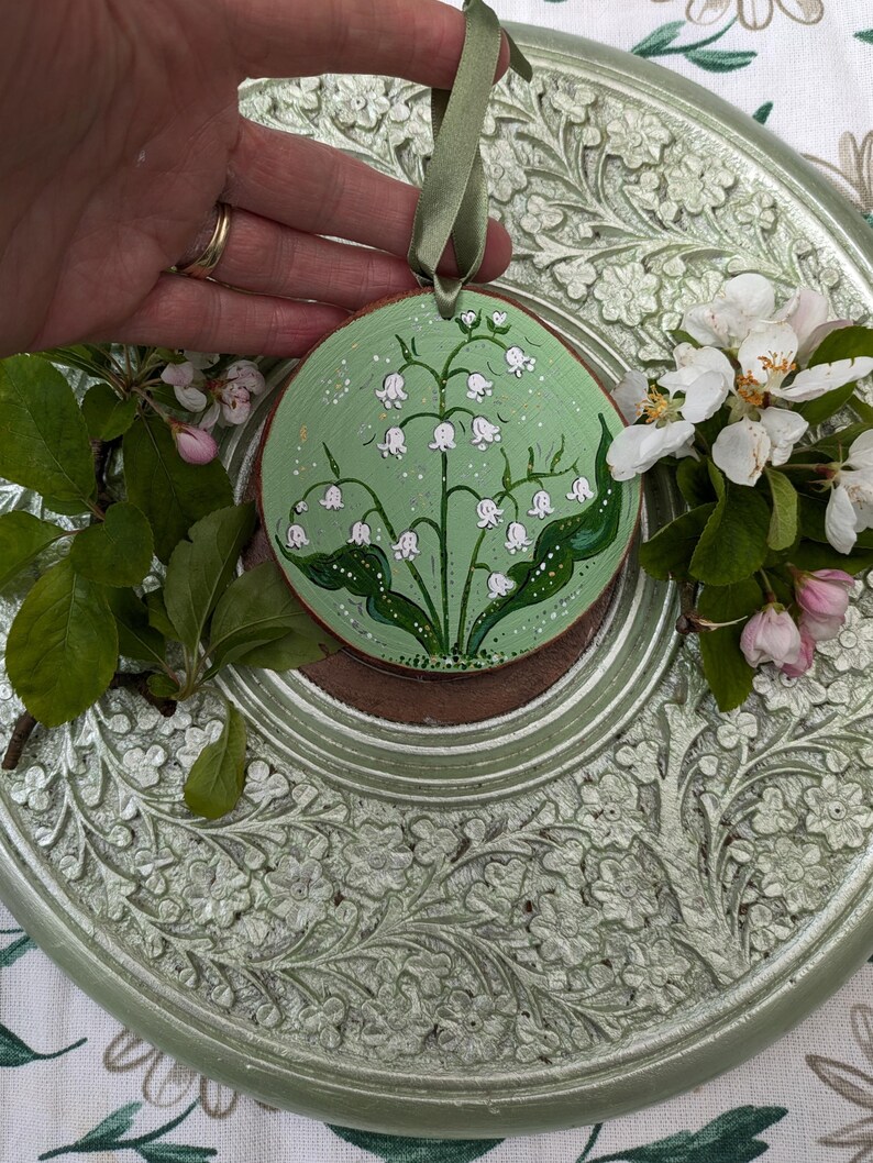May include: A round, wooden ornament with a hand-painted lily of the valley design. The ornament features a light green background with white flowers and green leaves. A green ribbon is attached for hanging. The ornament is approximately 4 inches in diameter.