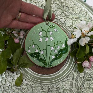 May include: A round, wooden ornament with a hand-painted lily of the valley design. The ornament has a light green background with white flowers and green leaves. A green ribbon is attached for hanging. The ornament is approximately 3 inches in diameter.