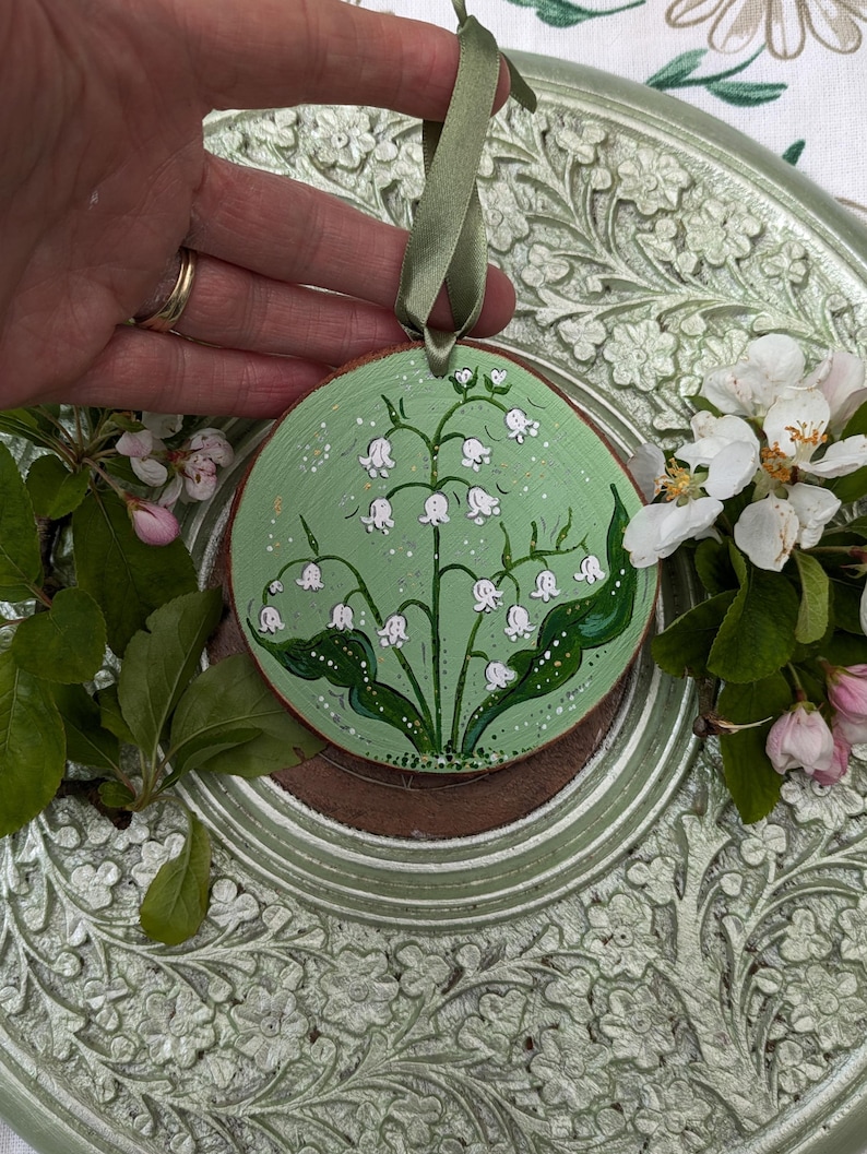 May include: A round, wooden ornament with a hand-painted lily of the valley design. The ornament has a light green background with white flowers and green leaves. A green ribbon is attached for hanging. The ornament is approximately 3 inches in diameter.