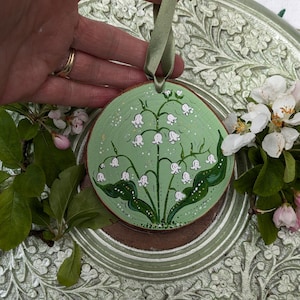 May include: A round, wooden ornament with a hand-painted lily of the valley design. The background is light green with white speckles. A green ribbon is attached for hanging. The ornament is approximately 4 inches in diameter.