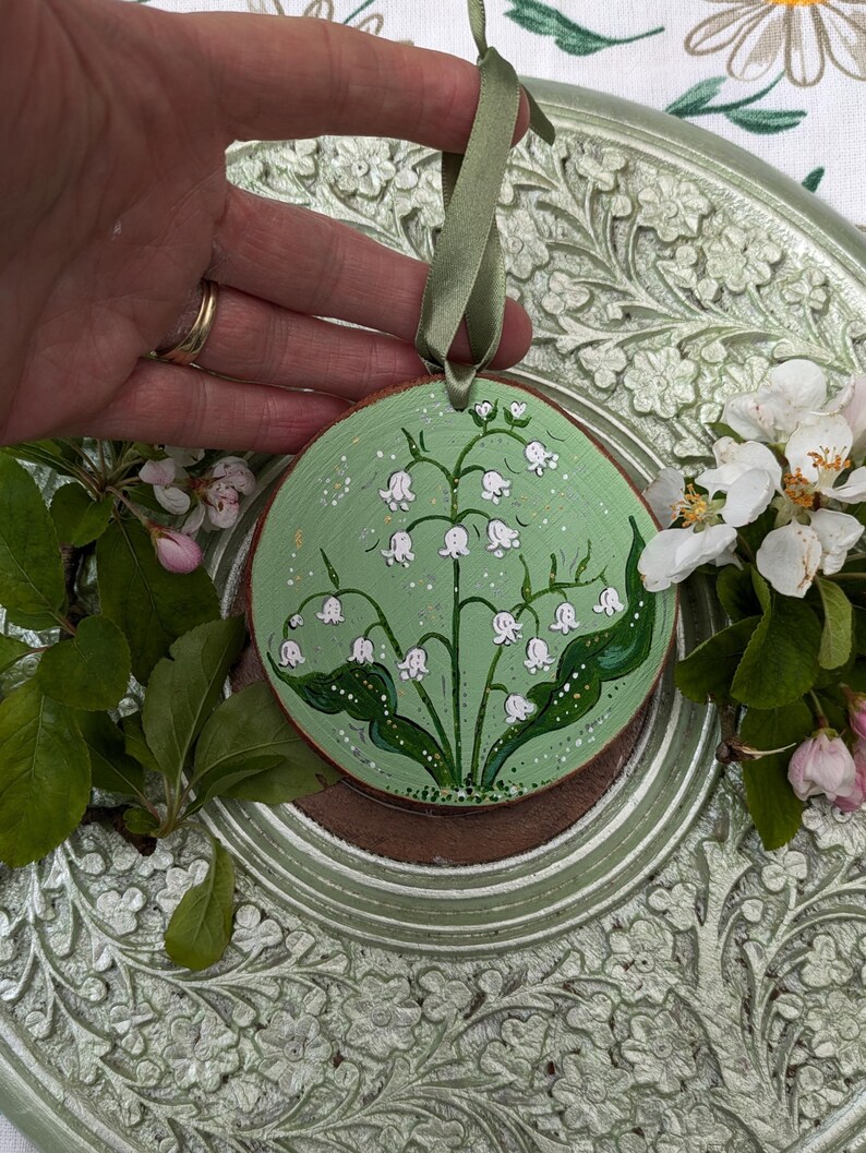 May include: A round, wooden ornament with a hand-painted lily of the valley design. The background is light green with white speckles. A green ribbon is attached for hanging. The ornament is approximately 4 inches in diameter.