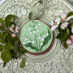 May include: A round, wooden ornament with a hand-painted lily of the valley design. The ornament is painted on a light green background and has a brown border. A green ribbon is attached for hanging. The ornament is surrounded by fresh flowers and leaves.